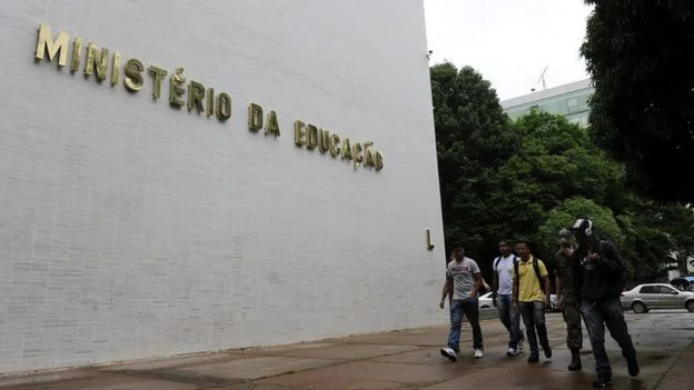 Fachada com letreiro do Ministério da Educação, na frente da qual passam cinco jovens caminhando