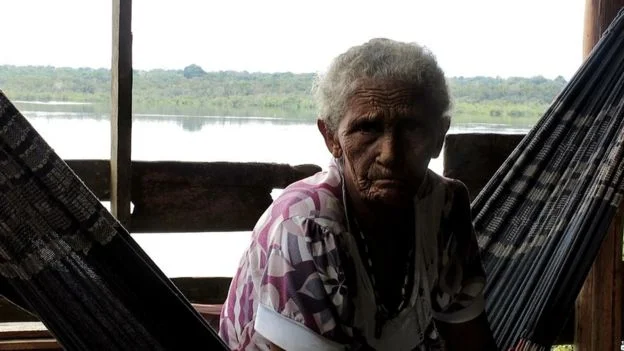 Idosa indígena sentada em uma rede ao lado de um rio em região próxima à estrada