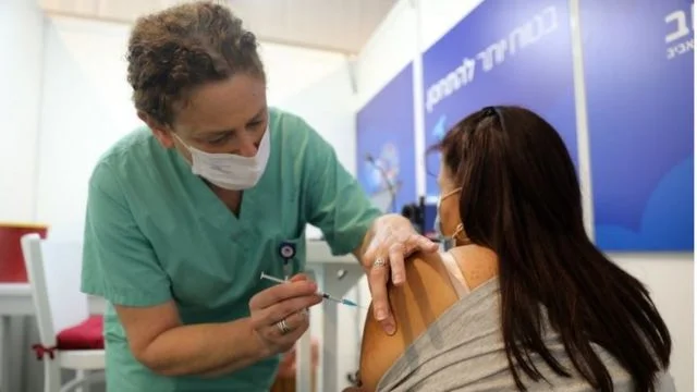 Dentro de sala, profissional aplica vacina em mulher de costas