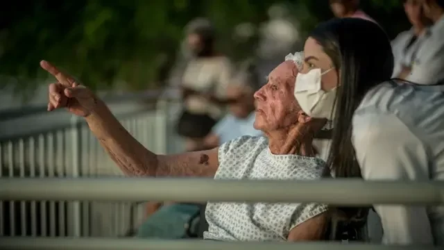 Foto de um senhor sentado apontado para a frente enquanto uma mulher de máscara e vestida de branco olha para onde ele aponta.
