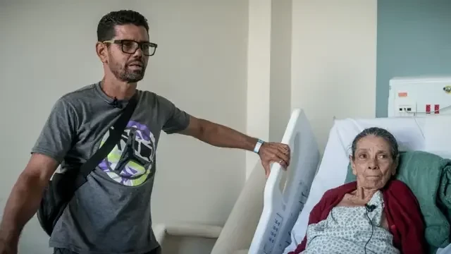 Foto de uma senhora em uma cama de hospital e, ao lado, de pé, um homem apoiado na cama. 