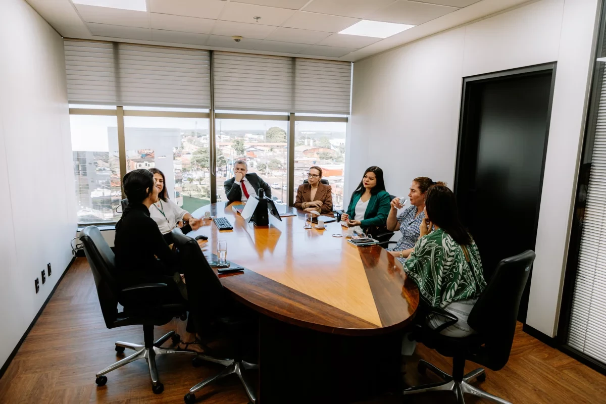 mesa de reunião com sete pessoas a mesa, dentre ela, um homem, todos vestidos formalmente, com janela ao fundo