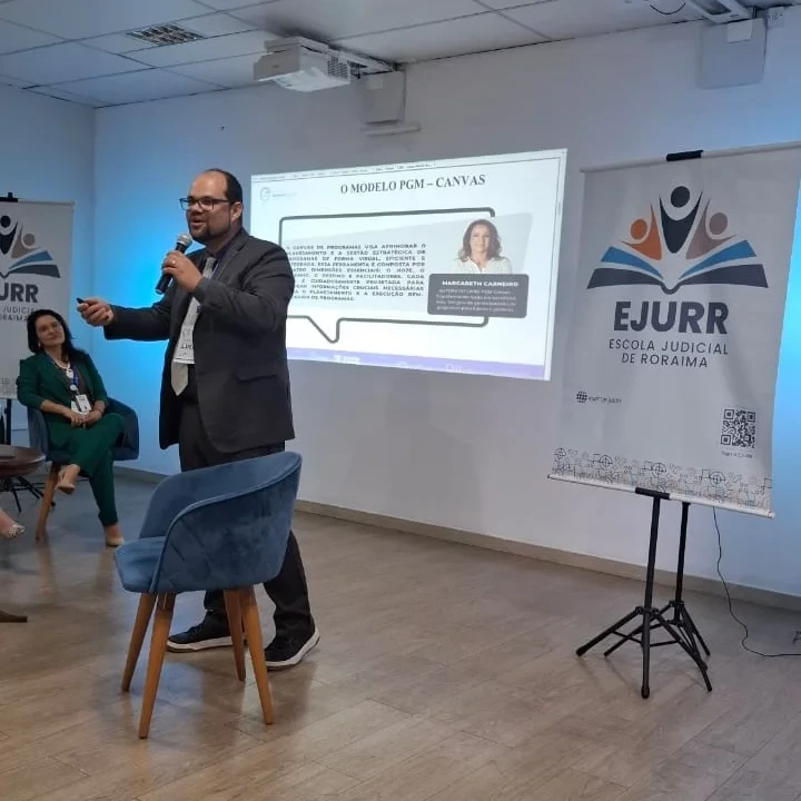 Foto de Lucas durante sua apresentação, à frente de uma projeção na parede, entre banners da Escola Judicial de Roraima; ele segura um microfone e utiliza crachá