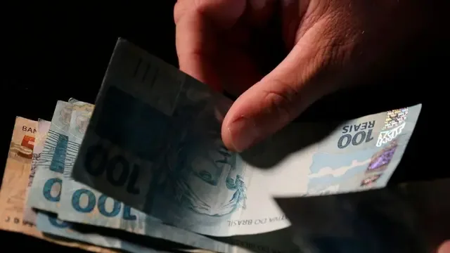 Mão segura nota de cem reais e a coloca sobre outras cédulas