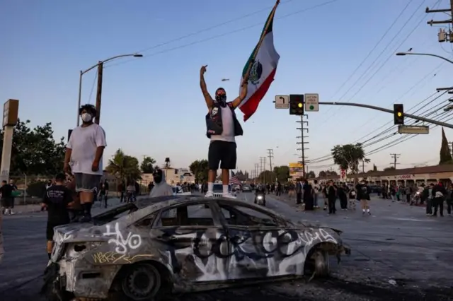 Manifestante com a bandeira do México em cima de um carro incendiado e pichado, em um protesto nas ruas