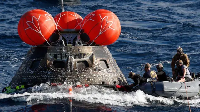 A cápsula metálica escura é vista flutuando na água, com três balões laranja inflados no topo e pessoal da Marinha fixando um cabo grosso em sua volta, em um bote salva-vidas