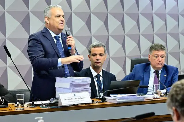 Gaspar fala ao microfone, de pé, com uma grande pilha de papéis a sua frente, seu relatório para a CPMI 