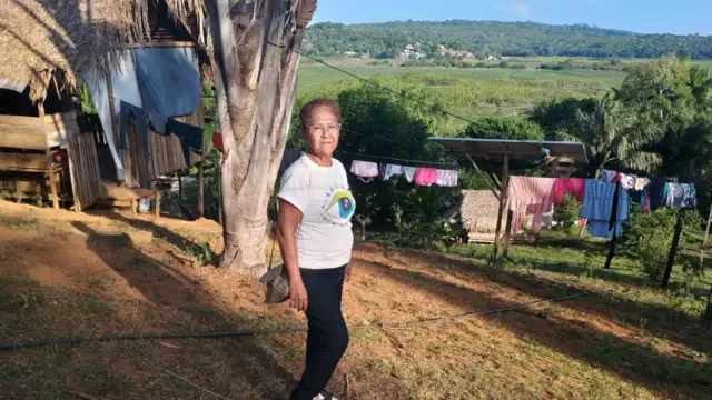 Uma mulher está em frente a uma casa simples de madeira, com roupas penduradas em um varal. Ao fundo, há uma área de vegetação e cultivo, com morros cobertos por floresta.