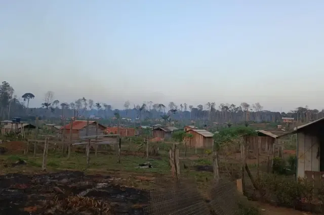 Área de ocupação rural na Amazônia, com casas simples de madeira espalhadas entre terrenos cultivados e cercas. Ao fundo, a vegetação foi amplamente desmatada, com árvores queimadas ou sem folhas e uma camada de fumaça sobre a paisagem.