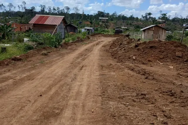 Estrada de terra em uma área de ocupação rural, cercada por casas simples de madeira e terrenos cultivados. Ao fundo, a floresta aparece parcialmente desmatada, com árvores derrubadas e vegetação espalhada pelo terreno.