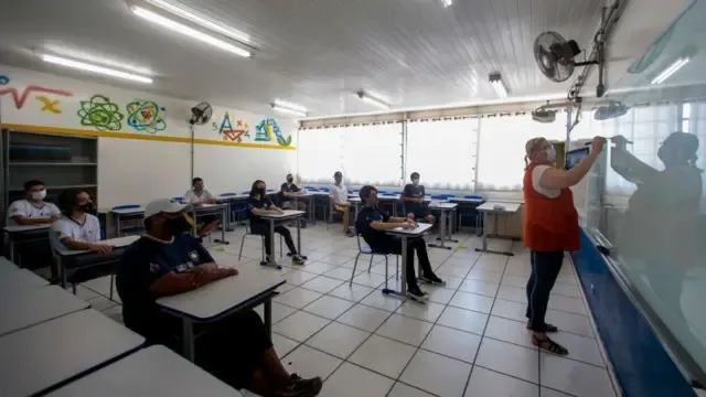 Uma professora escreve no quadro enquanto alunos, usando máscaras, acompanham a aula sentados em carteiras espaçadas em uma sala de aula. A cena mostra medidas de distanciamento adotadas durante a pandemia de Covid-19.
