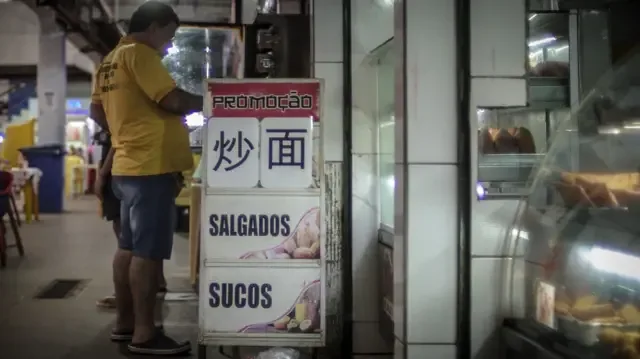 Imagem de lanchonete dentro do Mercado Central de Camaçari com foco em uma placa com os dizeres "Promoção, salgados, sucos, junto de ideogramas em mandarim".