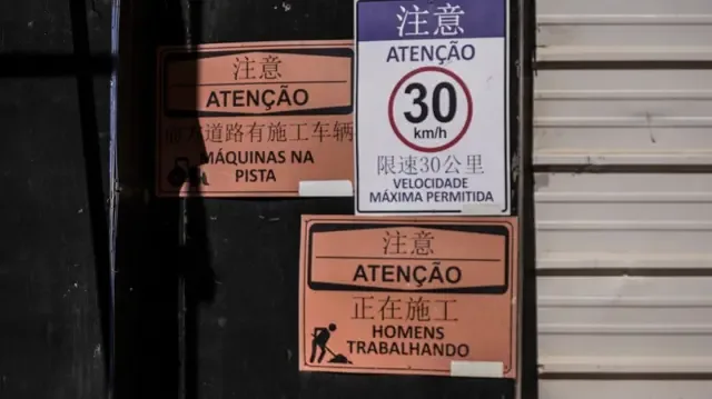 Entrada do canteiro de obras do alojamento da BYD com placas em português e mandarim com os dizeres "homens trabalhando", "máquinas na pista" e "velocidade máxima permitida 30km/h"