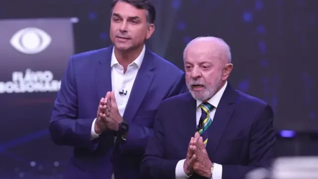 Flávio Bolsonaro e Lula em posições semelhantes, com mãos unidas em forma de oração, fotografados durante o debate nos estúdios da Band